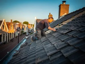 Dakdekker legt donkergrijze dakpannen op steil woonhuisdak in Nederlandse wijk onder gouden zomerzon.