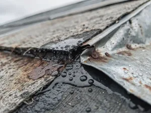 Verweerde daknok met gebarsten bitumenaansluiting, waterdruppels op vochtige dakbedekking en licht opgelichte zinkflits.