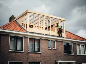 Dakopbouw in aanbouw op Nederlands rijtjeshuis met houten skelet, rode bakstenen gevel en dakdekker die constructie inspecteert.