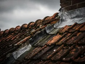 Verweerde dakrand met gebarsten voegwerk en aangetaste dakpannen bij schoorsteen, watervlekken op terracotta dakpannen onder bewolkte lucht.