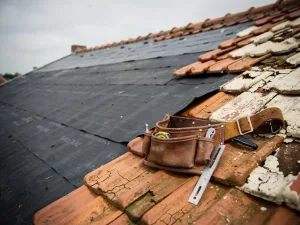 Verweerd dak in renovatie met vers bitumenmembraan naast gebarsten dakpannen en een leren gereedschapsriem op het dakoppervlak.