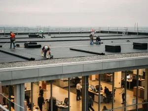 Dakdekkers installeren bitumineuze membraanplaten op plat commercieel dak terwijl bedrijfsactiviteiten binnen ongestoord doorgaan.