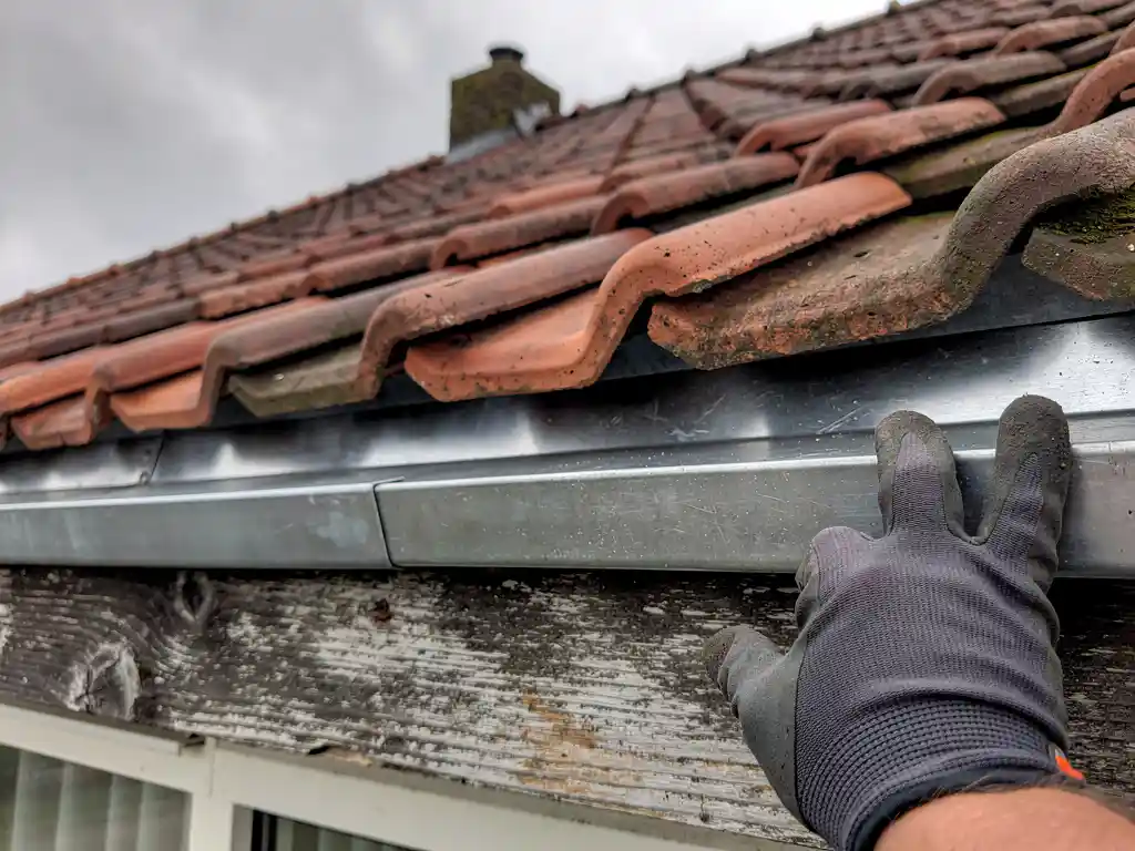 Dakdekker plaatst zinken dakrandafwerking langs de goot, met terracotta dakpannen en een bewolkte lucht op de achtergrond.