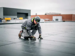 Dakspecialist inspecteert plat bitumineus dakmembraan van commercieel gebouw bij bewolkt daglicht.