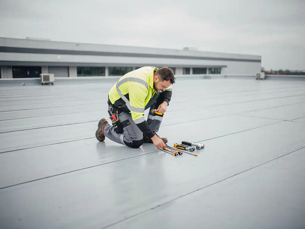 Dakinspecteur in gele veiligheidskleding knielt op plat commercieel dak en inspecteert membraanoppervlak met gereedschap.